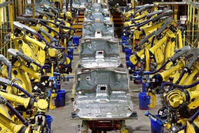 A row of bare aluminum Ford F-150 truck bodies moving down an assembly line, flanked by rows of yellow robotic welding arms inside a Ford manufacturing plant.