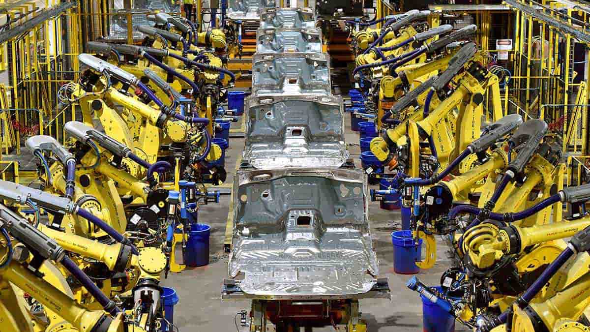 A row of bare aluminum Ford F-150 truck bodies moving down an assembly line, flanked by rows of yellow robotic welding arms inside a Ford manufacturing plant.