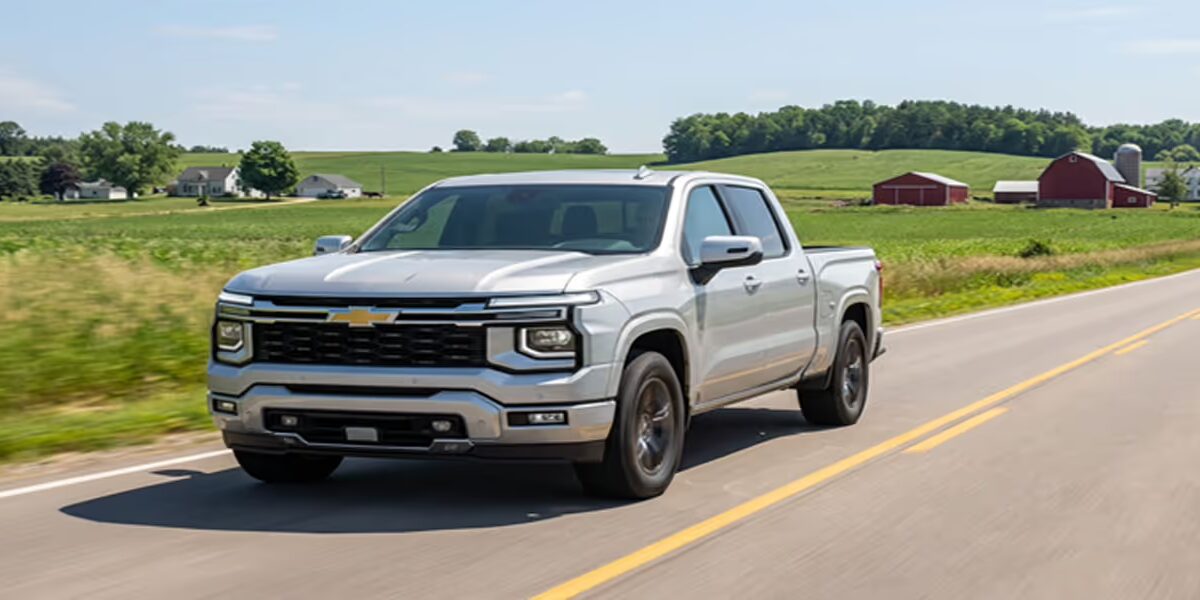 A silver 2027 Chevrolet Silverado crew cab pickup truck driving along a two-lane country road past green farmland and a red barn, photographed as a concept image.