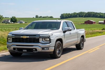 A silver 2027 Chevrolet Silverado crew cab pickup truck driving along a two-lane country road past green farmland and a red barn, photographed as a concept image.