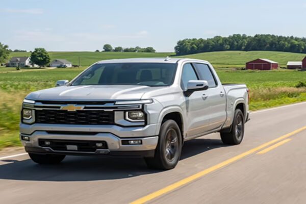 A silver 2027 Chevrolet Silverado crew cab pickup truck driving along a two-lane country road past green farmland and a red barn, photographed as a concept image.