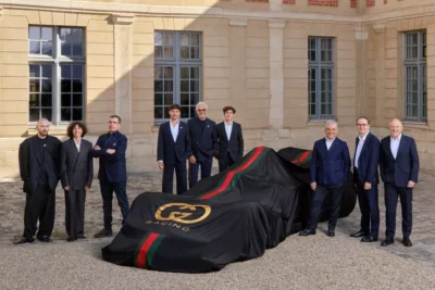 Gucci and Alpine F1 executives stand alongside a Formula 1 car draped in a black cover featuring the Gucci Racing logo and signature red and green stripes, photographed outside a French chateau at the partnership announcement.