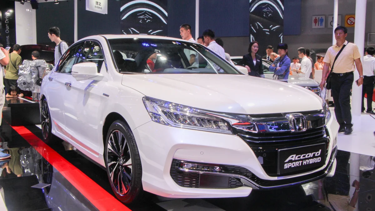 A white Honda Accord Sport Hybrid sedan on display at an auto show, with visitors browsing the stand and a cutaway hybrid powertrain visible in the background.