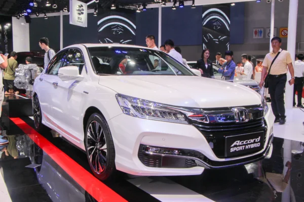 A white Honda Accord Sport Hybrid sedan on display at an auto show, with visitors browsing the stand and a cutaway hybrid powertrain visible in the background.