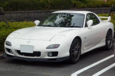 A white third-generation Mazda RX-7 FD with factory body kit and dark alloy wheels, parked in Japan, representing one of the final examples of the rotary-engined sports car that ended production in 2002