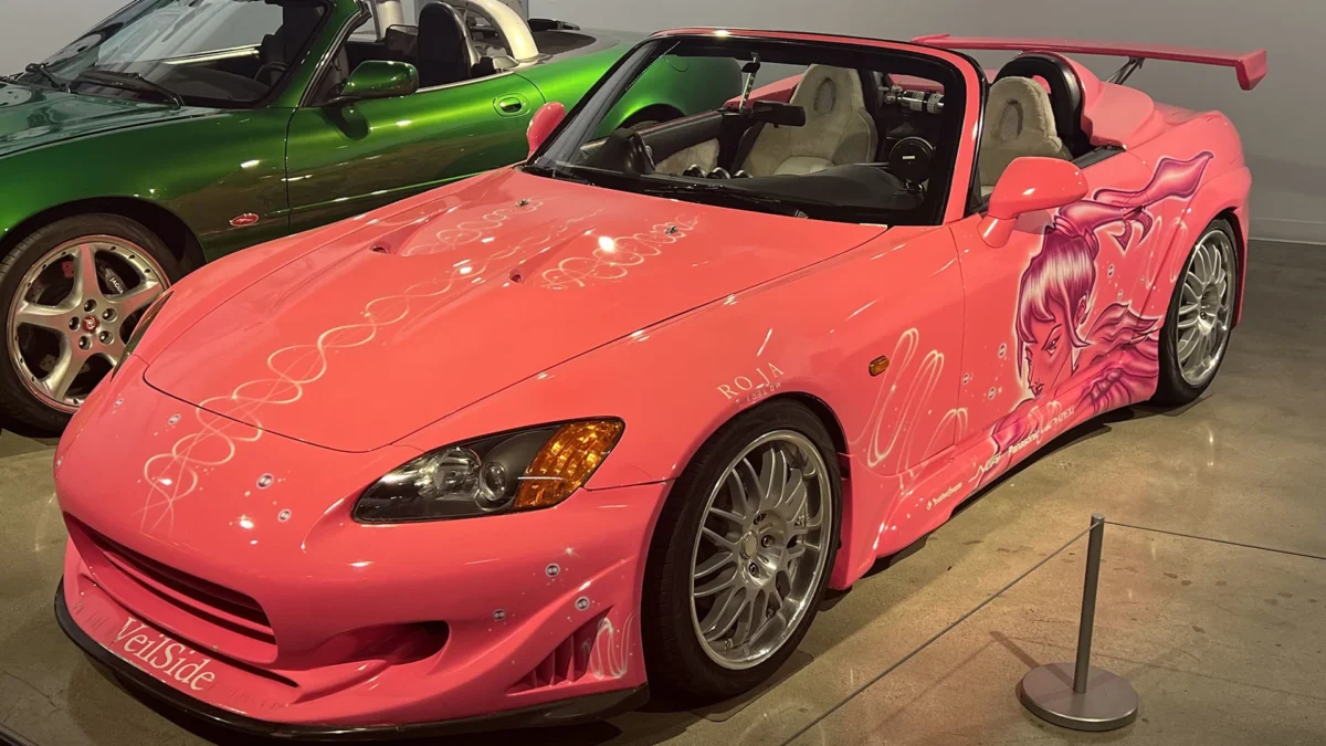 Suki's pink VeilSide-bodied 2001 Honda S2000 from 2 Fast 2 Furious, featuring custom anime-style artwork and a rear wing, on display at the Petersen Automotive Museum's Fast and Furious 25th anniversary exhibition, with a green sports car visible in the background.