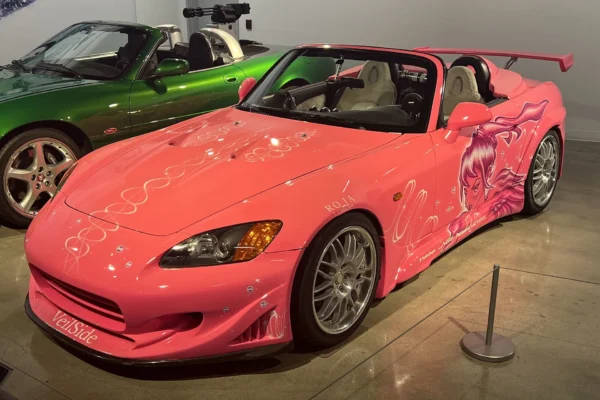 Suki's pink VeilSide-bodied 2001 Honda S2000 from 2 Fast 2 Furious, featuring custom anime-style artwork and a rear wing, on display at the Petersen Automotive Museum's Fast and Furious 25th anniversary exhibition, with a green sports car visible in the background.