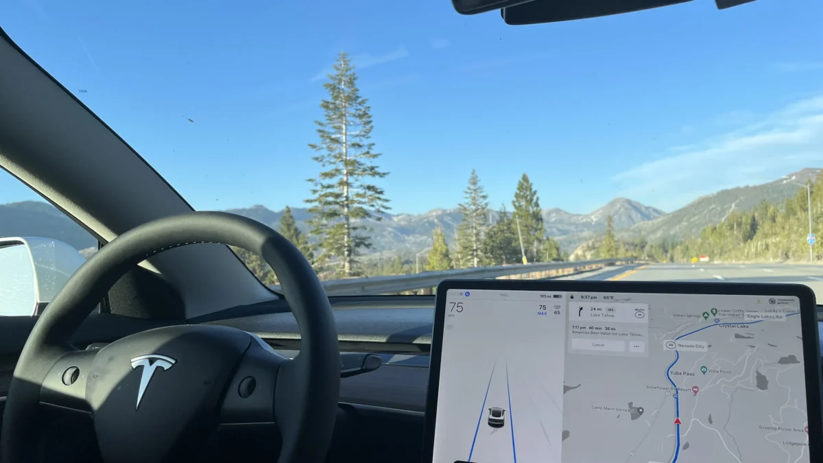 The interior of a Tesla Model 3 driving autonomously through a mountainous highway landscape, with the steering wheel untouched and the central touchscreen displaying FSD navigation guidance toward Lake Tahoe at 75mph.