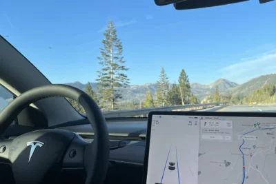 The interior of a Tesla Model 3 driving autonomously through a mountainous highway landscape, with the steering wheel untouched and the central touchscreen displaying FSD navigation guidance toward Lake Tahoe at 75mph.