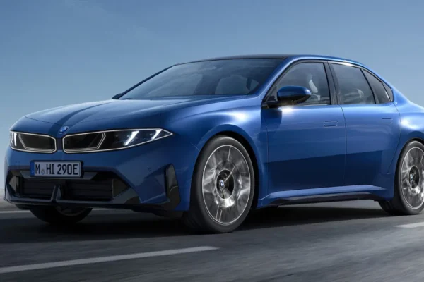 A Marina Bay Blue BMW i3 electric sedan photographed in dynamic three-quarter front view on an open road with a rocky mountain backdrop, showcasing the Neue Klasse platform's clean, minimalist exterior design with closed grille and aerodynamic alloy wheels.