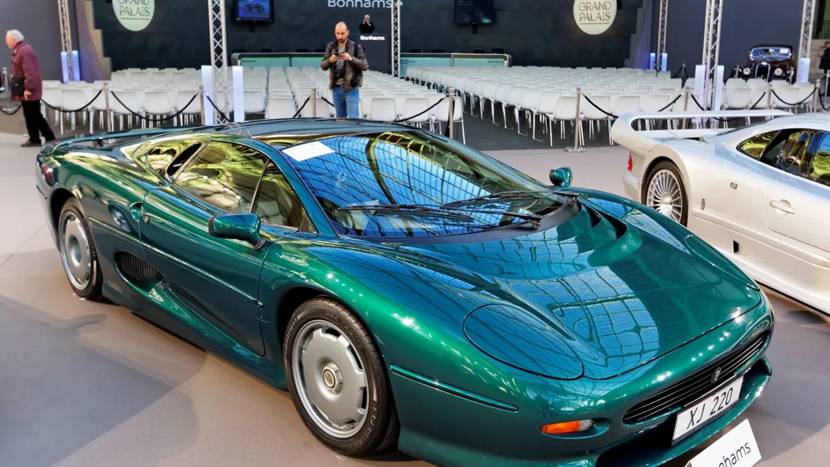 A teal-green Jaguar XJ220 supercar displayed at a Bonhams auction at the Grand Palais in Paris, photographed front-three-quarter with a white Mercedes CLK GTR partially visible in the background.