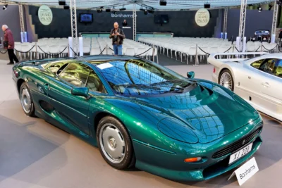 A teal-green Jaguar XJ220 supercar displayed at a Bonhams auction at the Grand Palais in Paris, photographed front-three-quarter with a white Mercedes CLK GTR partially visible in the background.