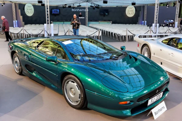 A teal-green Jaguar XJ220 supercar displayed at a Bonhams auction at the Grand Palais in Paris, photographed front-three-quarter with a white Mercedes CLK GTR partially visible in the background.