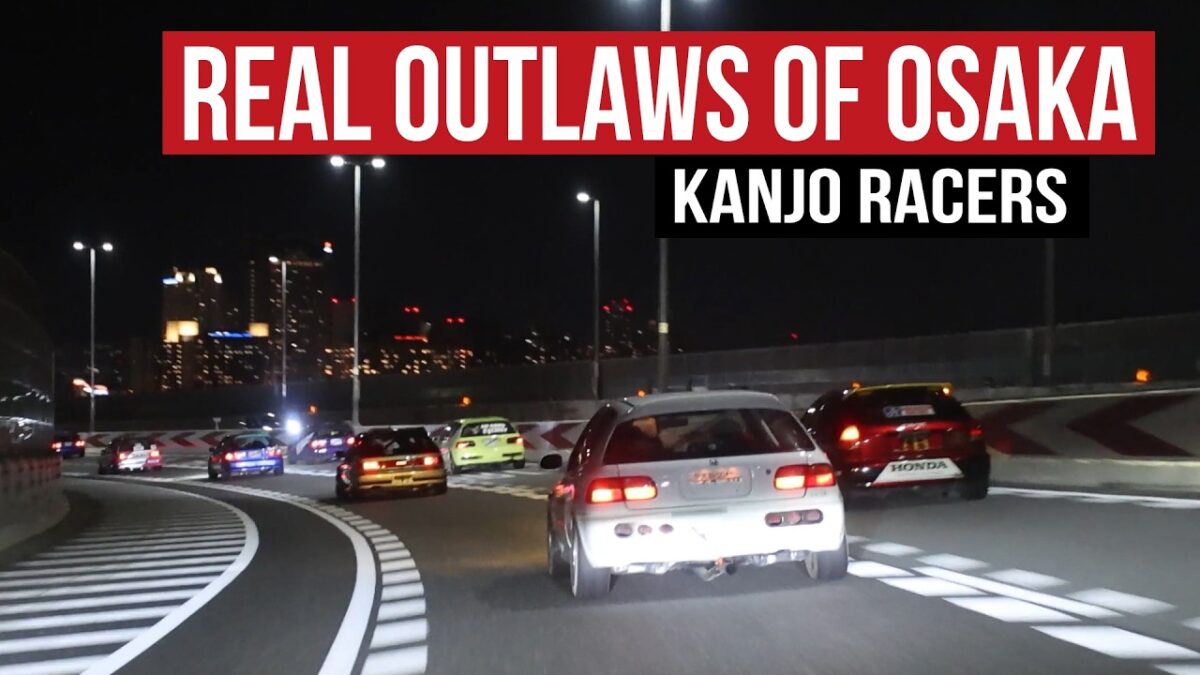 A pack of livery-covered Honda Civics running the Kanjo Loop on the Hanshin Expressway in Osaka at night, captured from behind as the No Good Racing Kanjozoku crew sweeps through a banked corner with the city skyline glowing in the background.