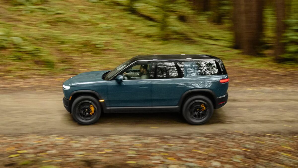 A teal Rivian R2 electric SUV photographed in dynamic side profile while driving along a forest trail, with motion blur on the background trees conveying speed and the vehicle's off-road capability.