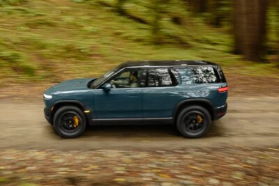 A teal Rivian R2 electric SUV photographed in dynamic side profile while driving along a forest trail, with motion blur on the background trees conveying speed and the vehicle's off-road capability.