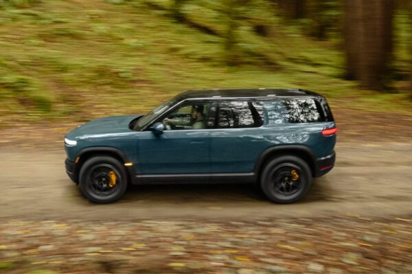 A teal Rivian R2 electric SUV photographed in dynamic side profile while driving along a forest trail, with motion blur on the background trees conveying speed and the vehicle's off-road capability.