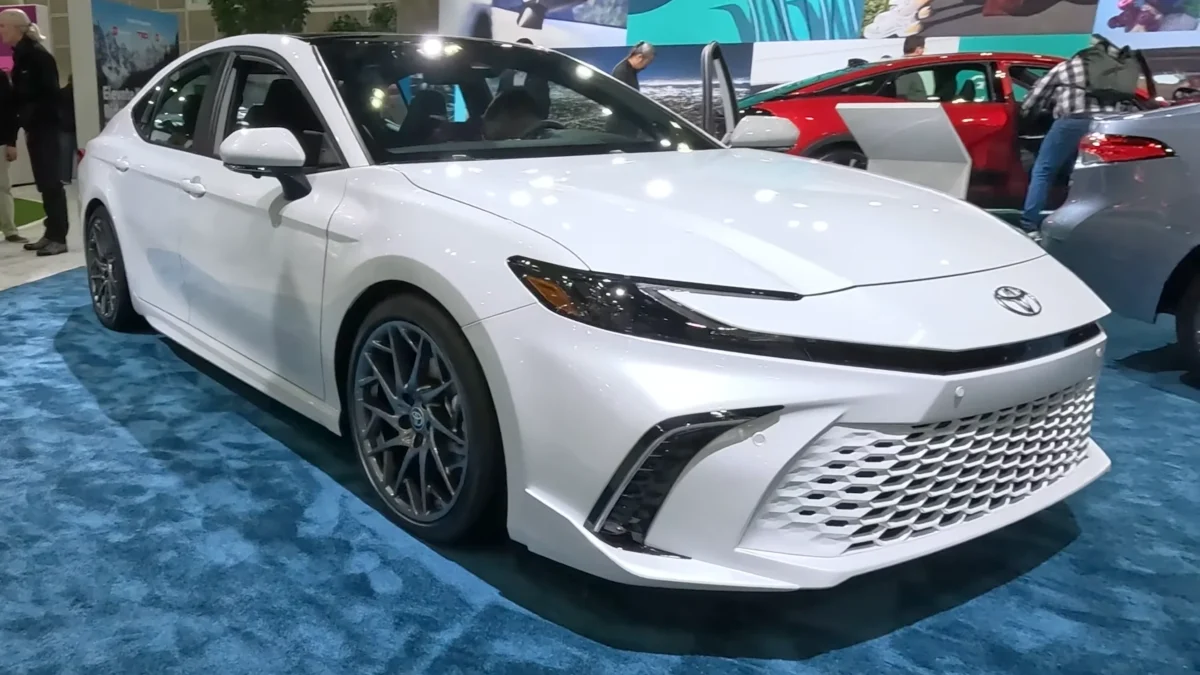 A white 2026 Toyota Camry Hybrid sedan on display at an auto show, photographed front-three-quarter with its sporty mesh grille and dark alloy wheels visible, with a red Camry visible in the background.