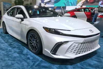 A white 2026 Toyota Camry Hybrid sedan on display at an auto show, photographed front-three-quarter with its sporty mesh grille and dark alloy wheels visible, with a red Camry visible in the background.