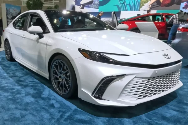 A white 2026 Toyota Camry Hybrid sedan on display at an auto show, photographed front-three-quarter with its sporty mesh grille and dark alloy wheels visible, with a red Camry visible in the background.