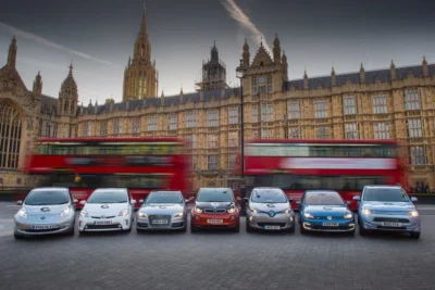 A lineup of seven early electric vehicles parked in front of the Houses of Parliament in London, including a Nissan Leaf, Toyota Prius Plug-in, Audi A3 e-tron, BMW i3, Renault Zoe, Volkswagen Golf GTE and Mitsubishi Outlander PHEV, with a red double-decker bus passing in motion blur behind them.