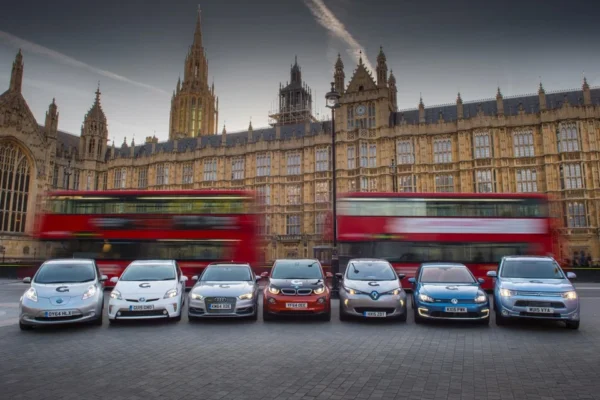 A lineup of seven early electric vehicles parked in front of the Houses of Parliament in London, including a Nissan Leaf, Toyota Prius Plug-in, Audi A3 e-tron, BMW i3, Renault Zoe, Volkswagen Golf GTE and Mitsubishi Outlander PHEV, with a red double-decker bus passing in motion blur behind them.