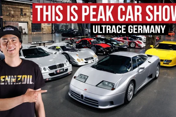Larry Chen stands in the industrial halls of Ultrace Germany 2026 at the Areal Böhler in Düsseldorf, pointing toward a collection of rare cars including a silver Bugatti EB110, a Mercedes-Benz CLK GTR street car, a yellow Ferrari F40, and several GT1-era Le Mans prototype race cars displayed on the polished concrete floor behind him.