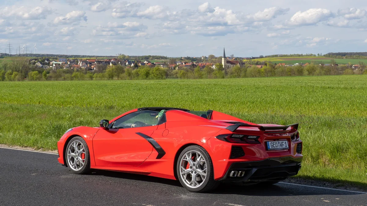 Red Chevrolet Corvette C8 convertible with top down, parked beside a green field with a German village and church spire in the background