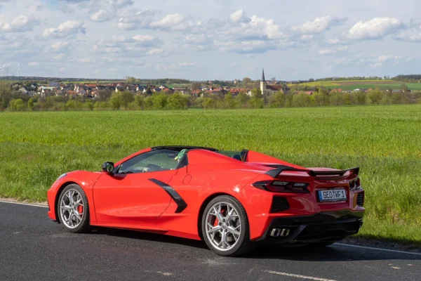 Red Chevrolet Corvette C8 convertible with top down, parked beside a green field with a German village and church spire in the background