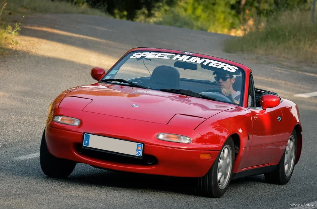 A first-generation Mazda MX-5 Miata NA in red on a winding road, representing the front-engine rear-drive layout that its chief engineer insisted upon over the mid-engine alternative